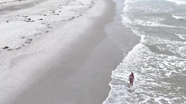 Mayo giymiş genç bir kadın kumlu bir plajda kıyı boyunca yürüyordu.