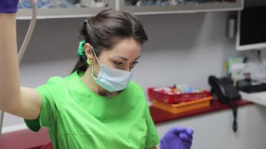 Yeşil üniformalı ve koruyucu maskeli bir dişçi klinikte diş aletleriyle çalışıyor.