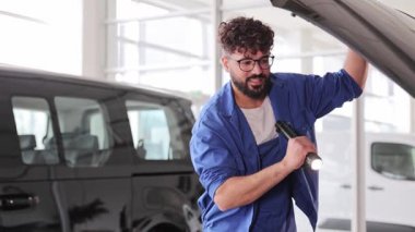 Mavi üniformalı genç araba tamircisi el feneriyle bir aracın motor bölümünü inceliyor.