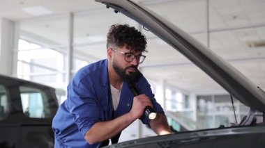 Mavi üniformalı profesyonel araba tamircisi el feneriyle araç motorunu inceliyor.