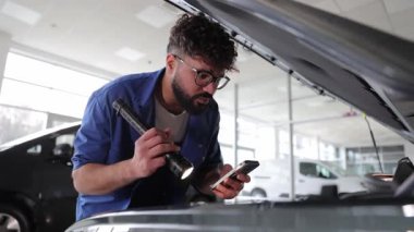 Garajda araç kontrolü yaparken akıllı telefon ve el feneri kullanan genç bir tamirci.