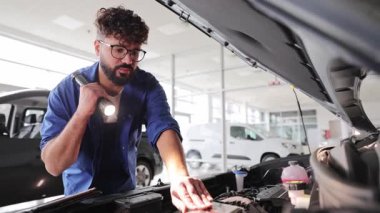 Profesyonel araba tamircisi bakım kontrolü sırasında el feneriyle araç motorunu inceliyor.