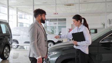 Genç bir satış elemanı, araba galerisinde potansiyel bir erkek alıcıya yeni bir araç sunuyor.