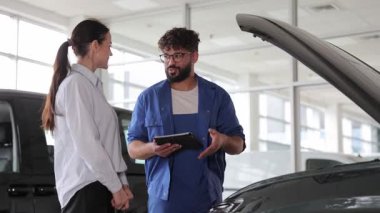Tabletli profesyonel araba tamircisi açık kaputlu bir arabanın yanında bayan bir müşteriye danışıyor.