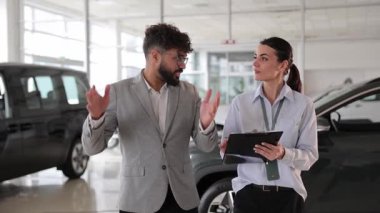Takım elbiseli bir adam modern bir galeride kadın satış danışmanıyla araba özelliklerini tartışıyor.