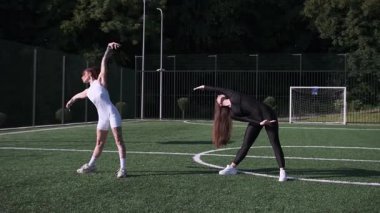 Yeşil bir spor sahasında spor yaparken eşofmanlı genç kadınlar birlikte esniyorlar.