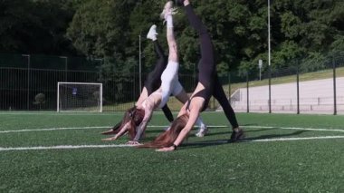 Spor kıyafetli genç kadınlar yeşil bir spor sahasında senkronize yoga pozları veriyorlar.