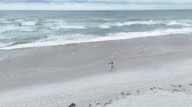 Geniş, boş bir sahil boyunca romantik gezen genç bir çiftin yüksek açılı görüntüsü.