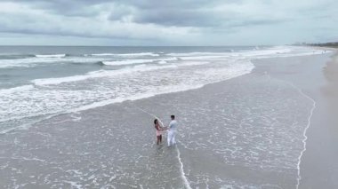 Bulutlu bir günde kumlu bir plajda birlikte yürüyen genç bir çiftin hava görüntüsü.