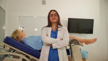 Genç bir jinekolog modern bir klinikte muayene koltuğuyla gülümsüyor.