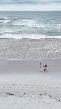 Genç bir çiftin el ele tutuşup okyanus kıyısında kumlu bir sahilde yürüdüğü yüksek açılı bir manzara.