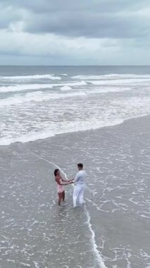 Genç mutlu ırklar arası çift, bulutlu bir günde el ele tutuşup sığ okyanus suyunda oynuyorlar.