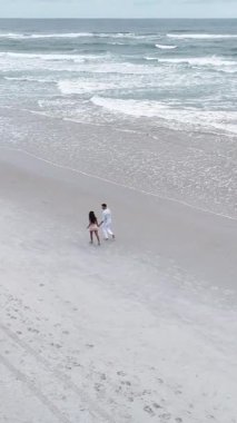 Genç bir çiftin el ele tutuşup güzel sahil şeridinde yürüdüğü yüksek açılı bir manzara.