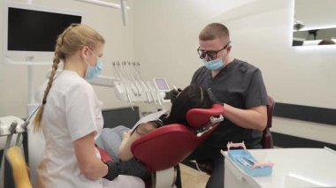 Profesyonel erkek dişçi ve asistanı dişçi koltuğundaki bir kadına diş tedavisi uyguluyor.