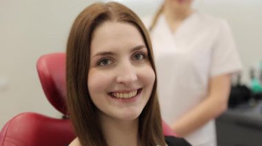 Arka planda bir dişçiyle dişçi koltuğunda otururken gülümseyen genç bir kadın.