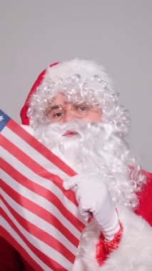 Noel Baba Amerikan bayrağını tutuyor ve gri arka plandaki kameraya bakıyor.