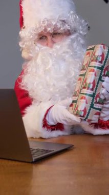 Uzun beyaz sakallı Noel Baba 'nın dizüstü bilgisayarında bir video görüşmesi var ve elinde bir hediye var.