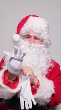 Noel Baba kostümlü bir adam gri arka planda beyaz eldivenlerini giyiyor.