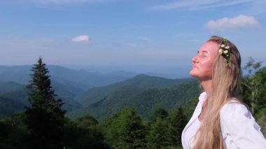 Saçlarında çiçeklerle vadiye bakan dağın tepesinde güneşin tadını çıkaran sakin bir kadın.