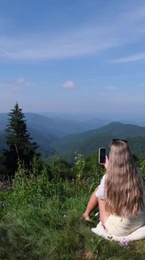 Çimenlerde oturan bir kadın güneşli bir günde güzel bir dağ vadisinin akıllı telefon fotoğraflarını çekiyor.
