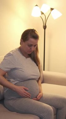 Beyaz tenli bir anne, bir lambanın yanındaki koltukta oturuyor ve elleri yuvarlak göbeğinde dinleniyor. Sıcak, yumuşak ışıklandırmalı ve kapalı mekanda huzurlu bir akşam atmosferi olan rahat bir doğum sahnesi..