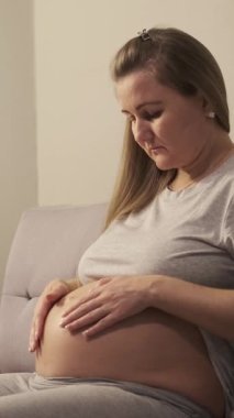 Hamile kadın evdeki kanepede otururken karnını nazikçe ovar. Dikey video.