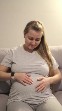 Hamile kadın evde rahat bir şekilde oturuyor, iki elini karnına koyuyor ve nazik bebek tekmelerine odaklanıyor..