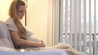 Hamile bir kadın evdeki koltukta oturuyor ve sıcak doğal ışıkta karnına hafifçe dokunuyor. Şefkat gösteren sakin bir annelik anı.