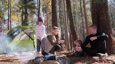 İki çocuklu mutlu bir aile çam ormanında kamp ateşinde marşmelov kızartıyorlar.