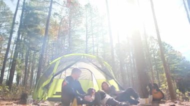 Genç bir çift güneşli bir çam ormanında çadırda dinlenirken köpeğini okşuyor.