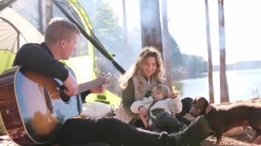 Çocuklu genç bir aile ve kamp yapan bir köpek, göl kenarında gitarla şarkı söylüyorlar.