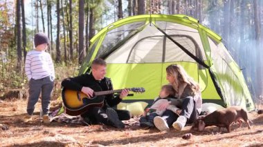 İki çocuğu ve bir köpeği olan beyaz bir aile piknik yapıyor ve ormanda bir çadırın yanında gitar çalıyorlar.