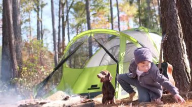 Genç çocuk ve köpeği ormanda sigara içilen bir kamp ateşinde birlikte oynuyorlar.