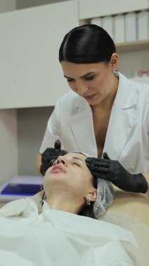 Profesyonel güzellik uzmanı güzellik kliniğinde bir kadının yüzüne kozmetik kömür maskesi takıyor.