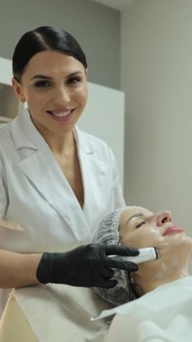 Güzellik salonunda bir kadının yüzünde mikro akım cihazı kullanarak gülümseyen bir kozmetikçi.