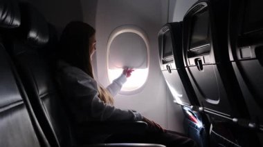Genç bayan yolcu güneş ışığının içeri girmesine izin vermek için bir uçağın camını açıyor.