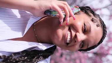 Parkta akıllı telefonuyla konuşurken saçları örgülü neşeli Afrikalı Amerikalı kadın gülümsüyor.