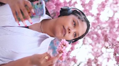 Kulaklıklı genç bir kadın pembe kiraz çiçekleriyle dolu bir parkta telefonuyla selfie çekiyor.