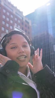 Kulaklıklı neşeli genç bir kadın güneş ışığı ve şehir binalarıyla dışarıda şarkı söylüyor ve dans ediyor.