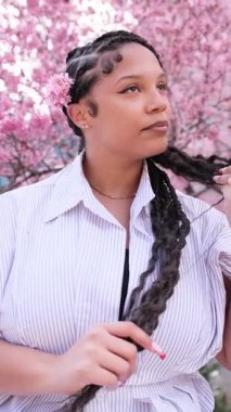 Saçları örgülü genç bir kadın, çiçek açan pembe kiraz ağaçlarıyla bir parkta bahar gününün tadını çıkarıyor.