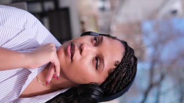 Saçları örgülü, kulaklıkla müzik dinleyen, dışarıda kameraya bakan genç siyahi bir kadın.