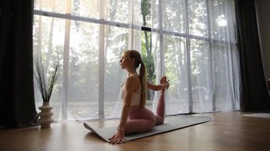 Genç atletik kadın büyük bir pencerenin önündeki mindere yoga pozu veriyor.
