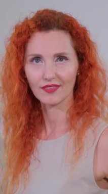 Beyaz bir arka plana gülümseyen kızıl kıvırcık saçlı muhteşem olgun bir kadının portresi.