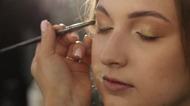 Profesyonel makyöz, genç bir kadının göz kapağına kozmetik fırçasıyla göz farı sürüyor.