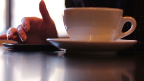Молода жінка сидить біля вікна в кафе, використовуючи смартфон і п'є каву — стокове відео