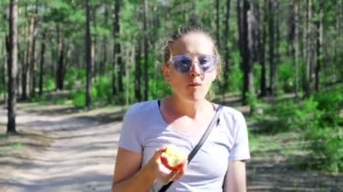 Güneş gözlüklü bir kız ormanda yürür ve yaz günü elma yer.