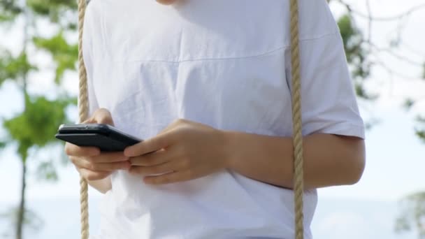 Meisje in zonnebril maakt gebruik van een smartphone terwijl zitten op een touw swing — Stockvideo