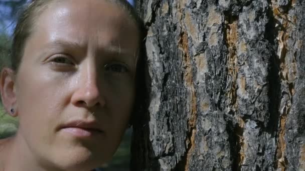La chica mira profundamente a la cámara, inclinando su cabeza contra un árbol — Vídeo de stock