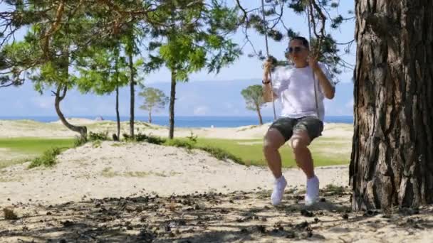 A girl swings on a rope swing under a tree on a sandy beach — Stock Video