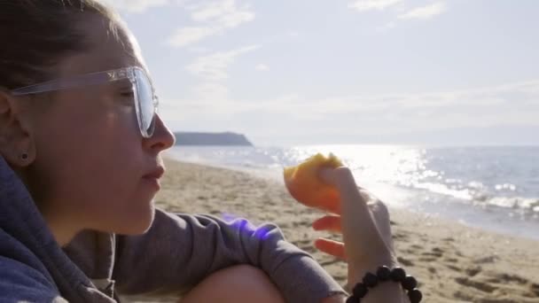 Girl sitting on the beach near the water and eating an apple — Stock Video